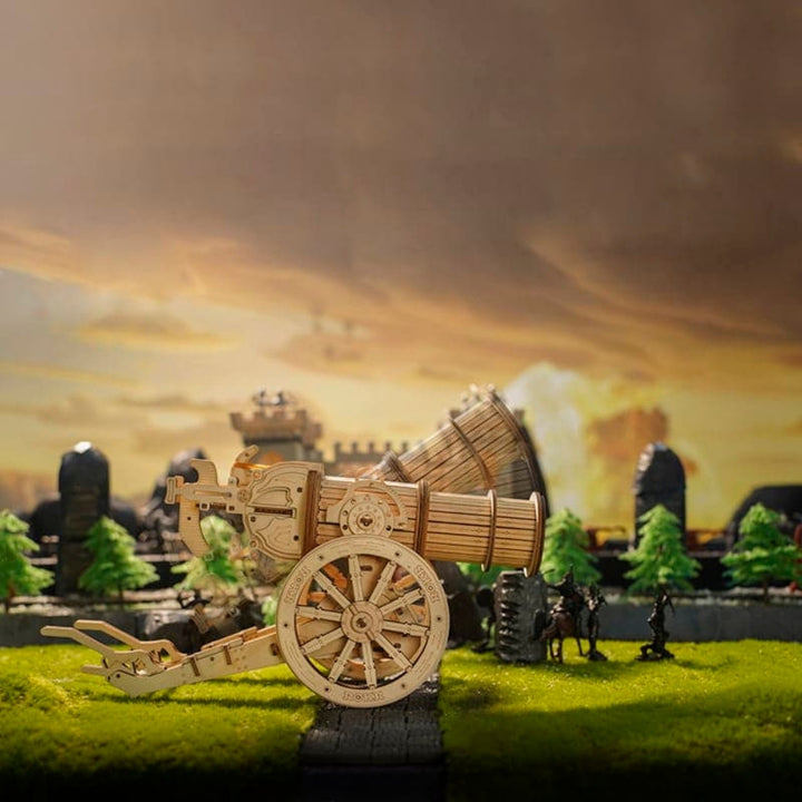 Wooden model of a cannon on a grassy field with a castle and trees in the background.